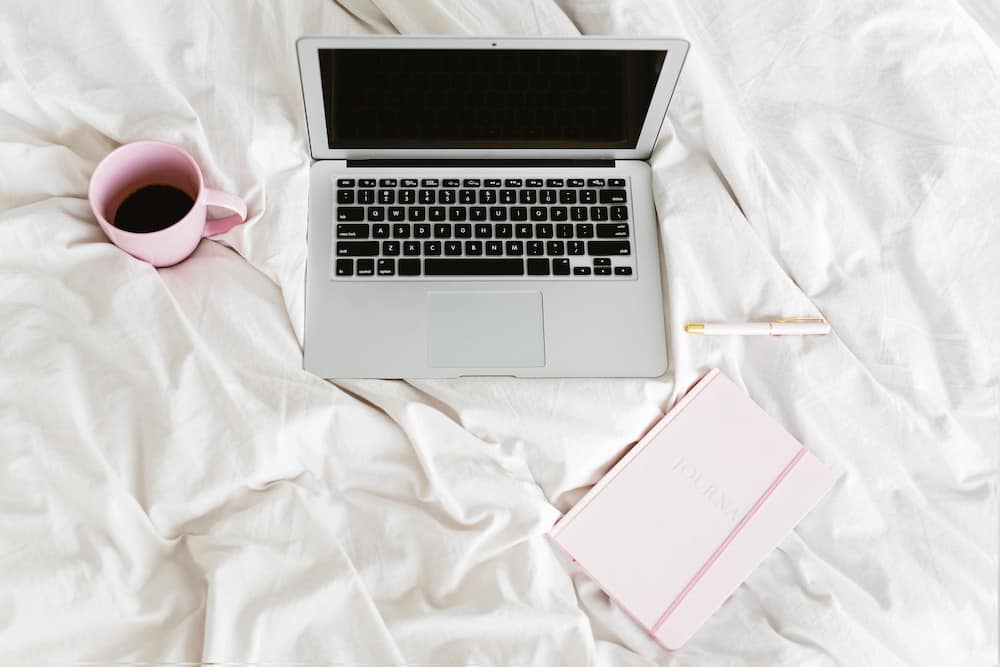 Silver laptop on a white bed with pink coffee cup and pink notebook