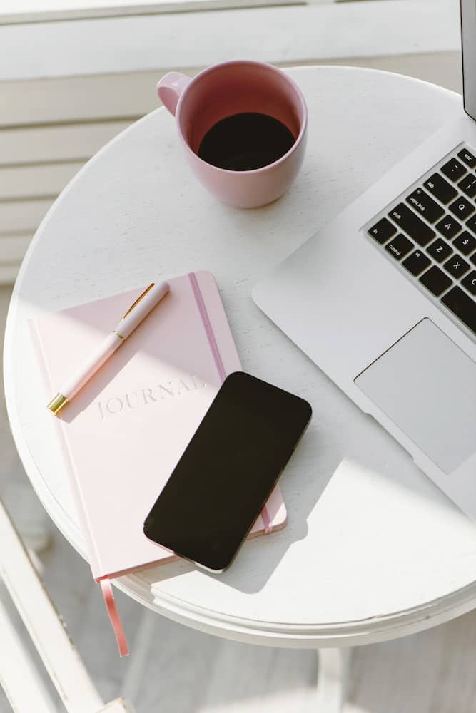 Phone on a pink journal beside a coffee cup and laptop. 