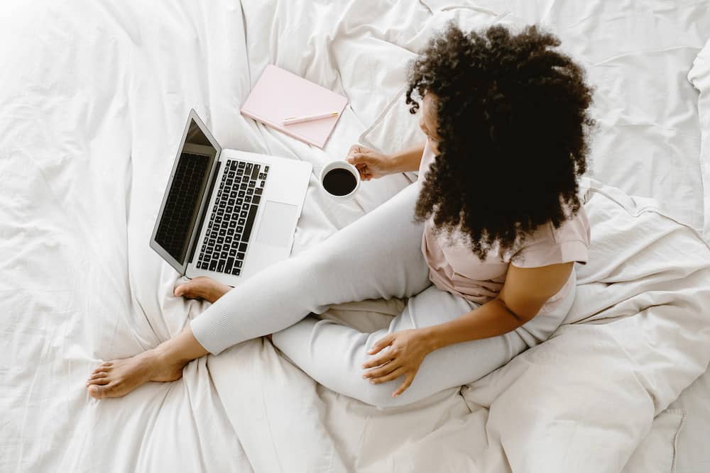 Black woman on a bed surrounded by coffee, her pink notebook and her laptop to work on her blog