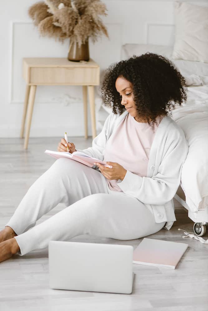 Black woman writing in a pink journal beside her laptop planning her blog.