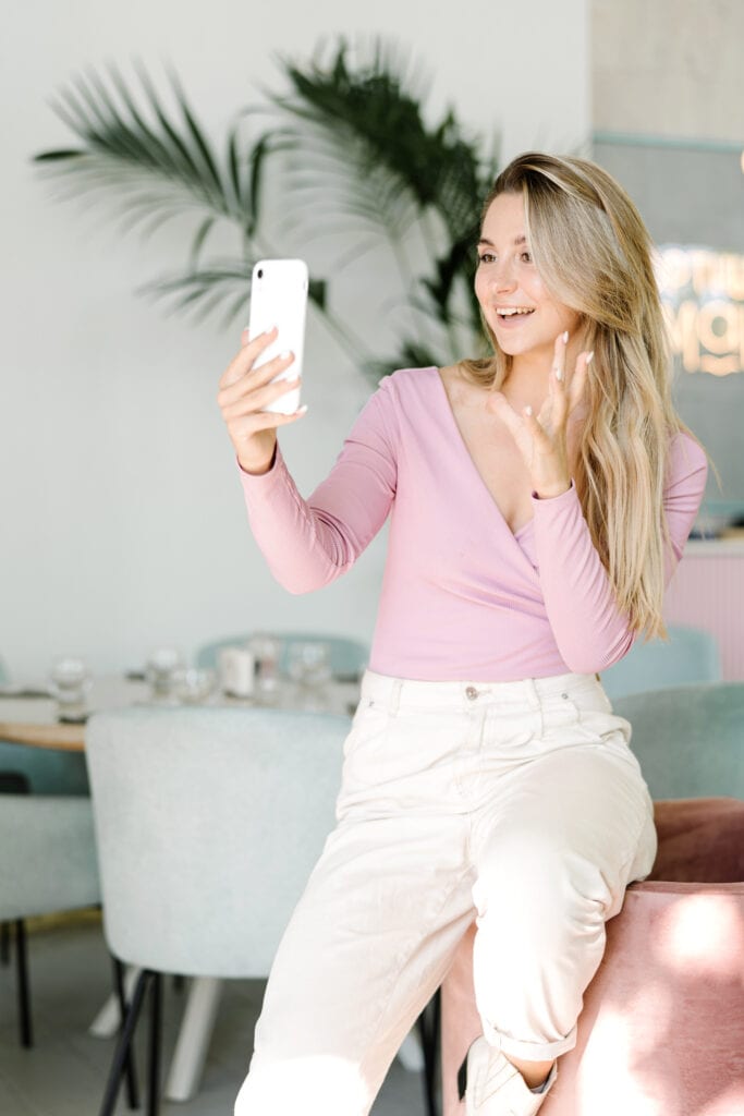 Blonde woman recording a video on her iPhone for her blog. She will upload it to her social media and her Youtube channel. She is standing in her living room gesturing with her hands towards the camera. 