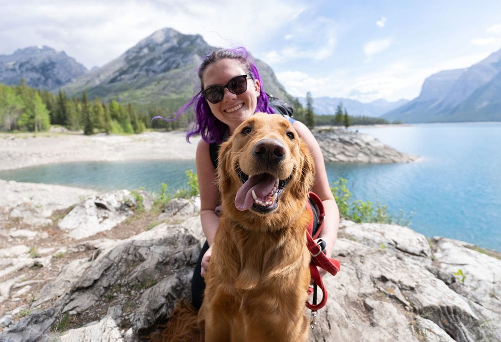 Nina, with purple hair and sunglasses, sits with her happy golden retriever, Theo, on a rocky trail by a crystal-clear lake, surrounded by mountains and trees in Banff National Park. This Moraine Lake hike is one of the best for dogs in Banff, and a great photo stop.